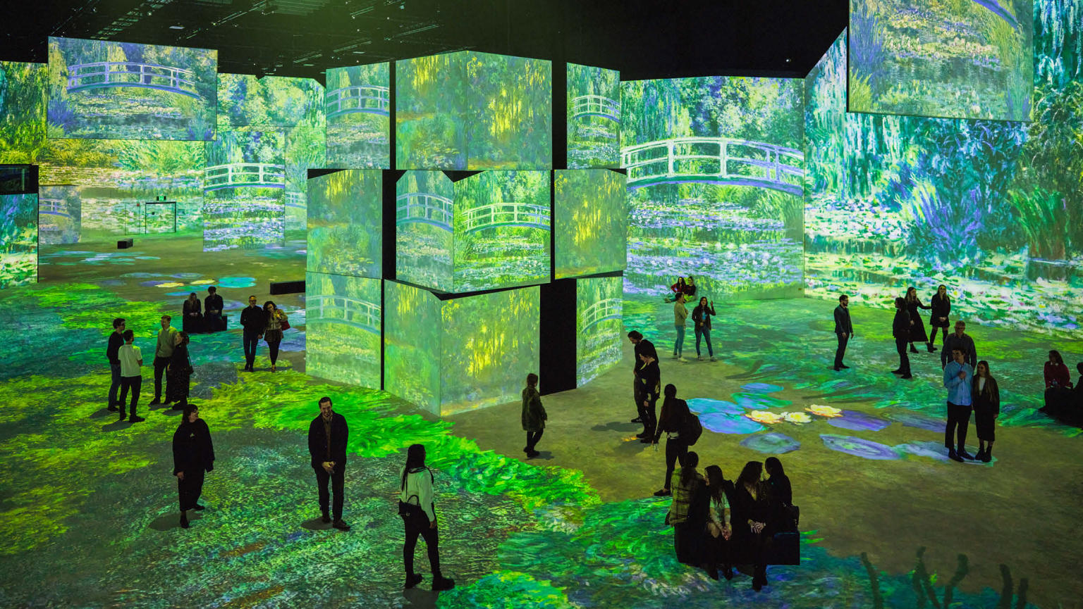 A group of individuals scattered around a spacious room adorned with an array of digital screens, including a central pillar of screens. The walls and screens showcase captivating artwork featuring Monet's renowned masterpiece, 'Bridge over a Pond of Water Lilies'. The immersive display transports viewers into the serene world of Monet's iconic creation.