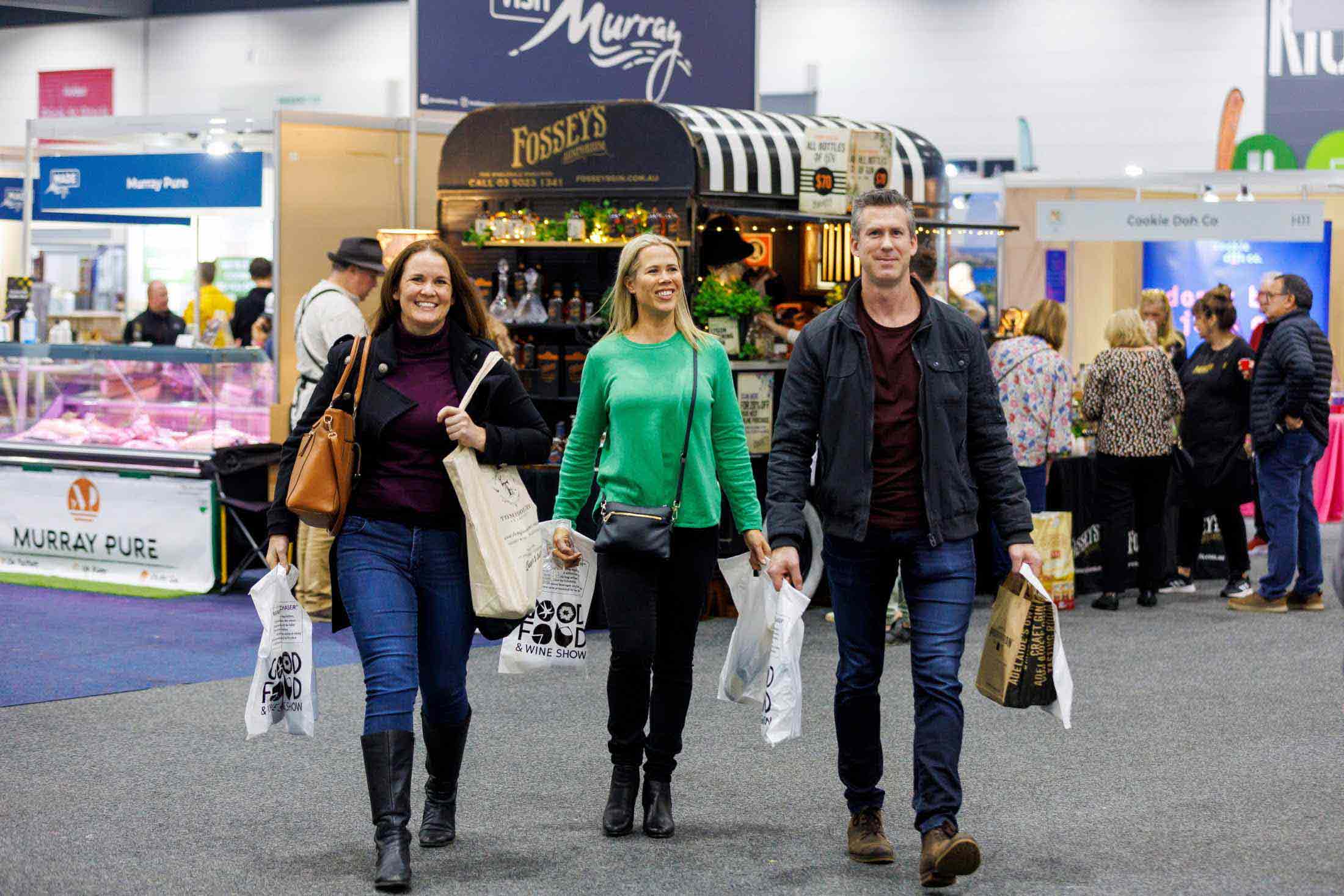 Three people walk towards the camera with lots of bags in hand. Exhibition stands and a crowd is seen in the background. 