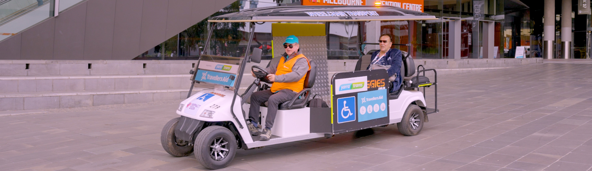 A driver in a high-vis orange vest driving a travellers aid cart with a passenger in the back outside MCEC. 