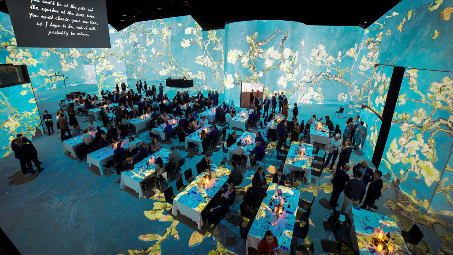 A large group of people seated at long rectangular tables within a spacious room enveloped by surrounding digital screens. The screens display a captivating artwork featuring a serene blue background adorned with elegant white flowers, creating an immersive ambience.