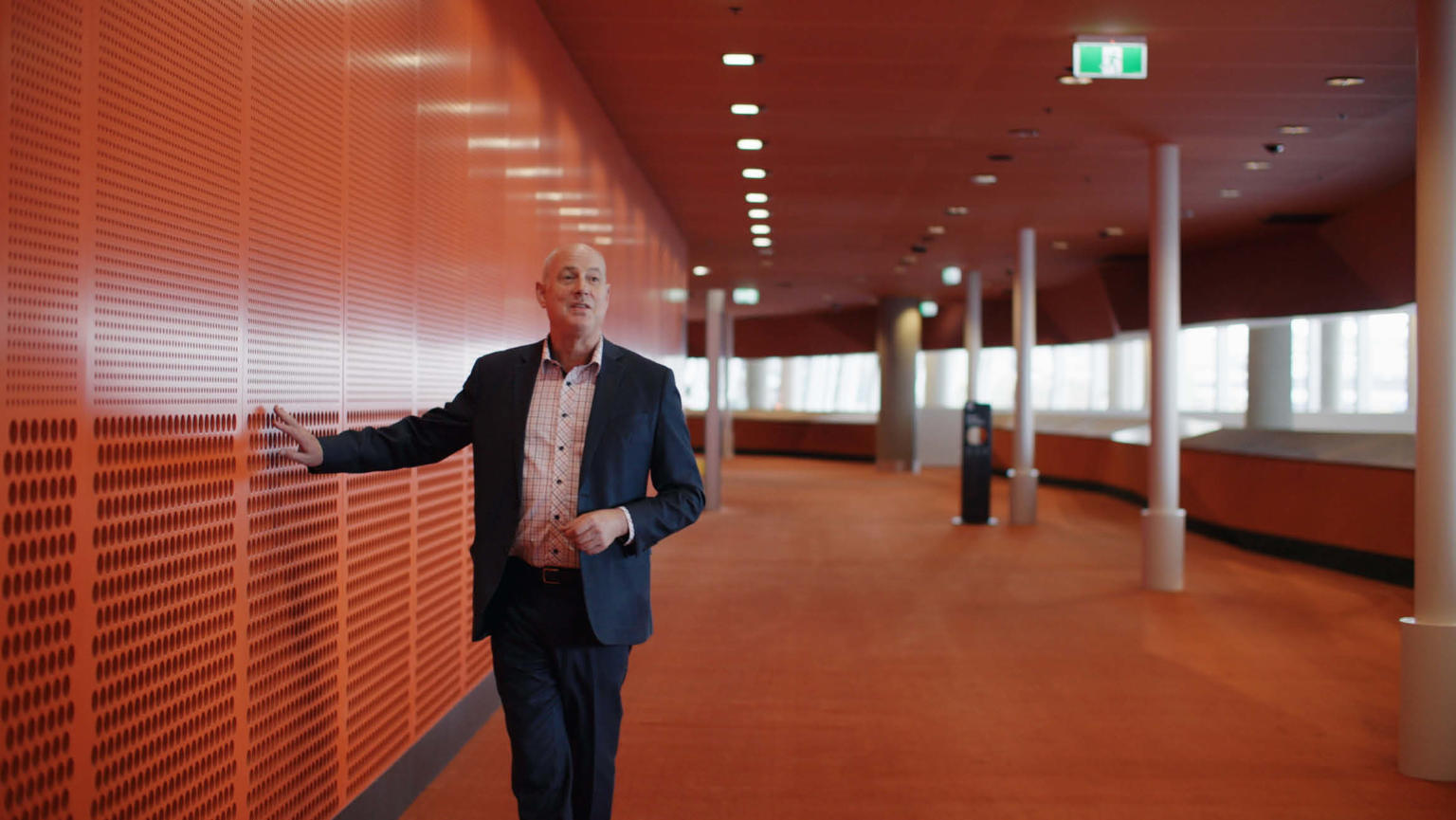 David Howie, a man in a suit, walking along the foyer in the MCEC (Melbourne Convention and Exhibition Centre) building. He is seen touching the orange wall.