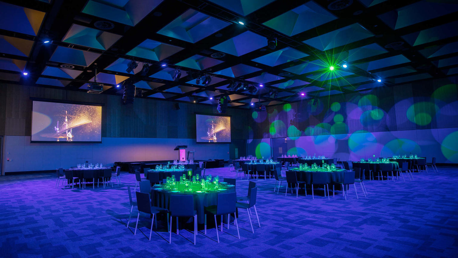 Empty room of distanced round tables, set for a gala dinner. The room is filled with blue lights and green spotlights on each table. 