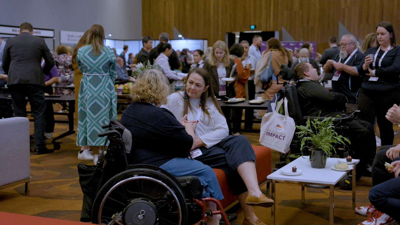 Two women are chatting in a busy convention. One lady is in a wheelchair while the other is sitting on an orange couch. 