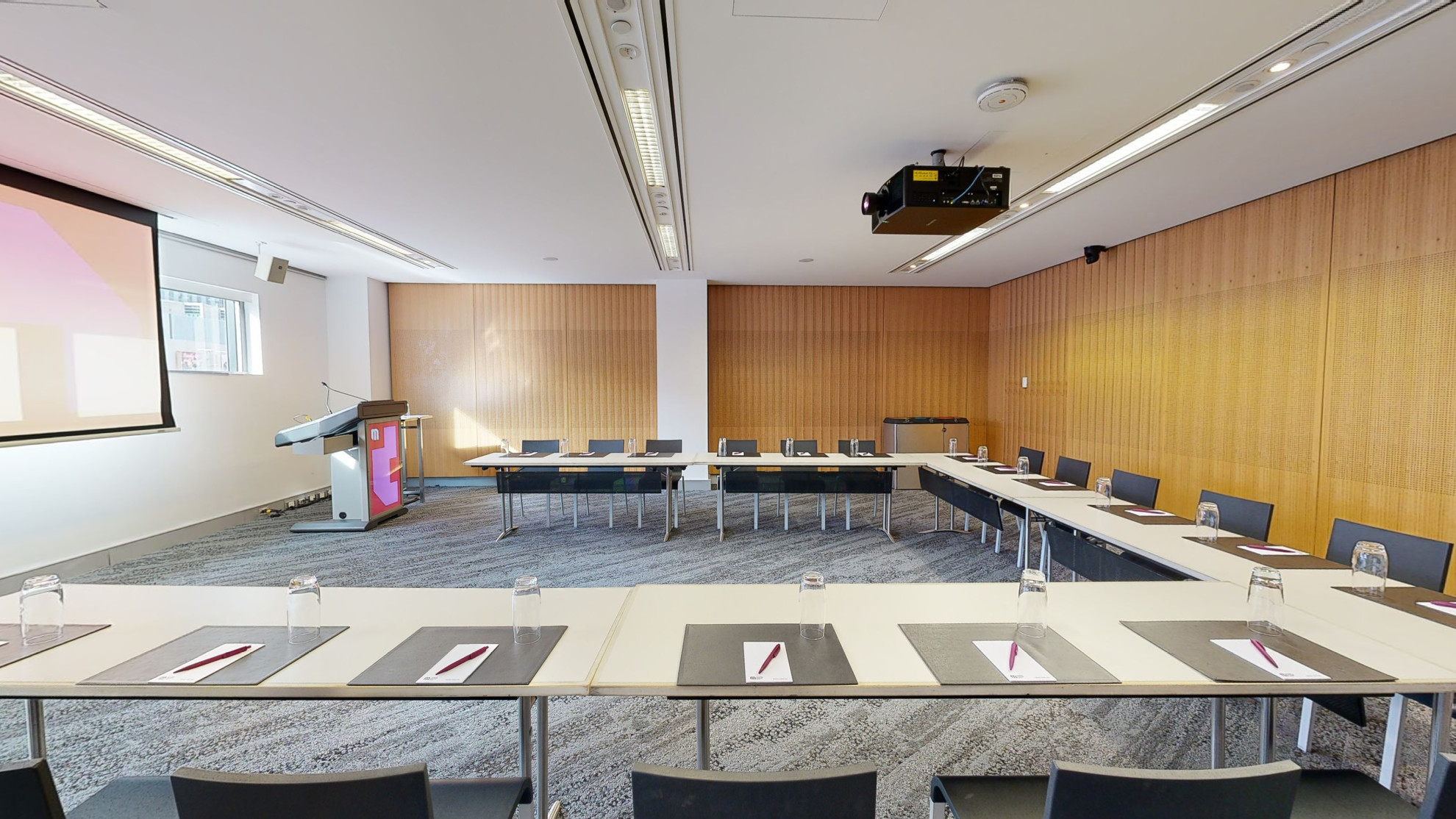 A meeting or conference room with tables and chairs arranged in a u-shaped boardroom configuration. The tables and chairs are facing a large projector screen and lectern both which have MCEC branding displayed on it. 