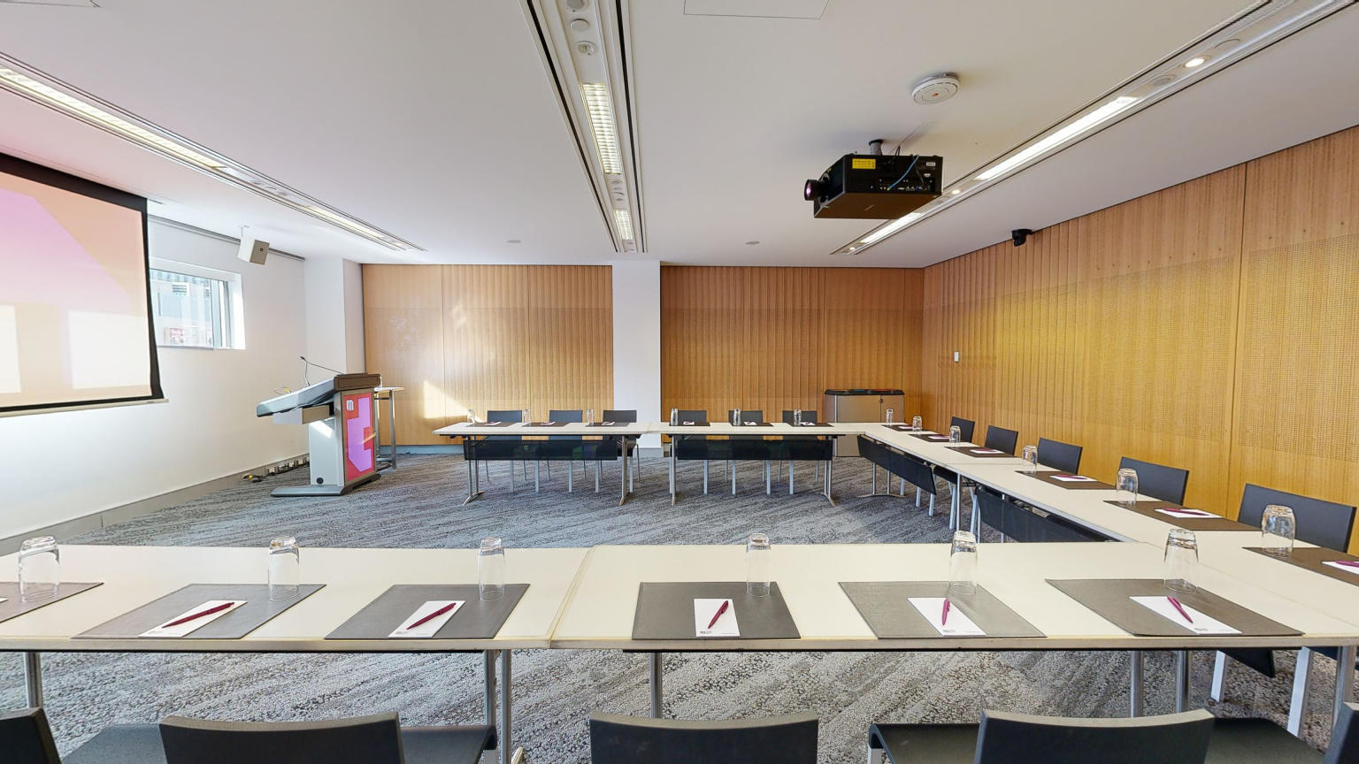 A meeting or conference room with tables and chairs arranged in a u-shaped boardroom configuration. The tables and chairs are facing a large projector screen and lectern both which have MCEC branding displayed on it. 