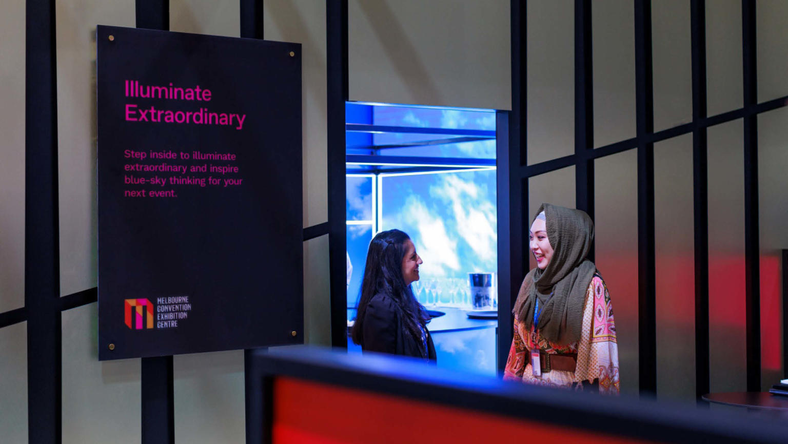 An image showing two women talking and smiling at each other behind a red and grey exhibition stand with a digital sign next to them reading 'Illuminate Extraordinary'. 