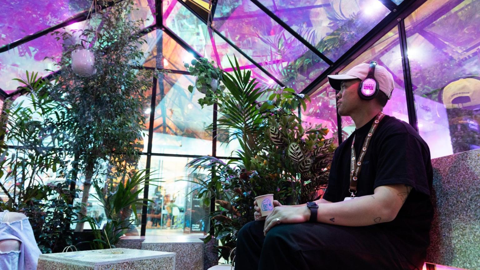 A man sitting with headphones on in a colour glass house with plants around. 