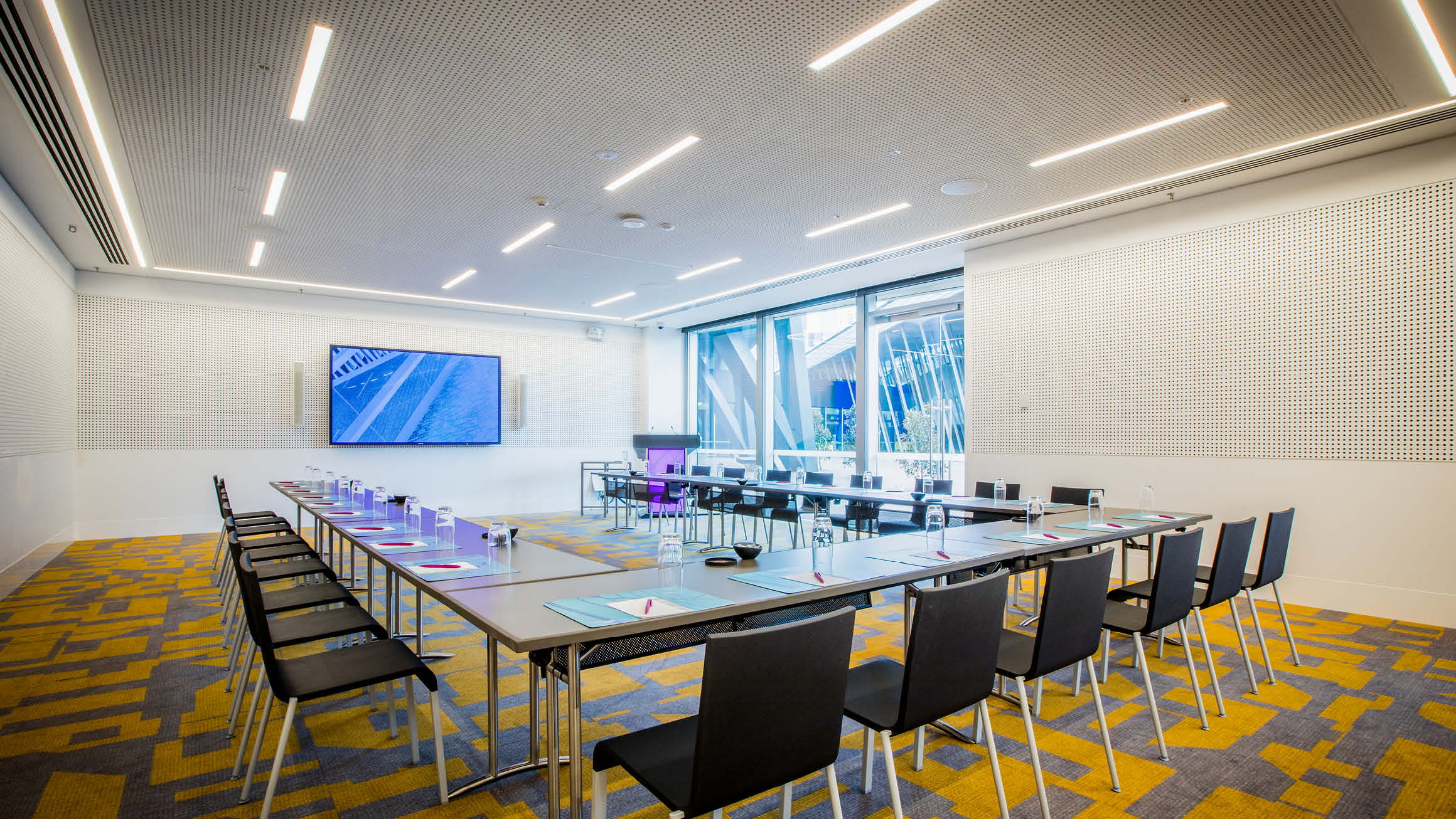 Empty room of table and chairs set up in a u-shaped boardroom. The chairs face a screen at the front of the room, Windows are situated in the far corner letting in some natural light. 