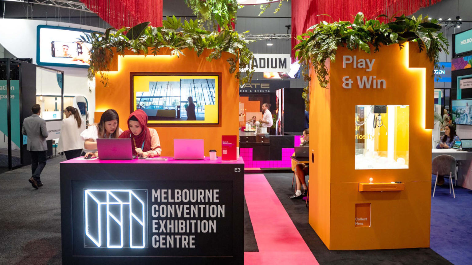An attention-grabbing exhibition stand with a dynamic arrangement of elements: long red fringing hanging from the ceiling, a vibrant orange wall featuring a walk-through cutout. On the left side, a pink and black table proudly displays a neon logo reading 'Melbourne Convention and Exhibition Centre' with two women standing behind it looking at a laptop screen. To the right, a captivating claw drop game beckons with the enticing words 'Play & Win' above it. 