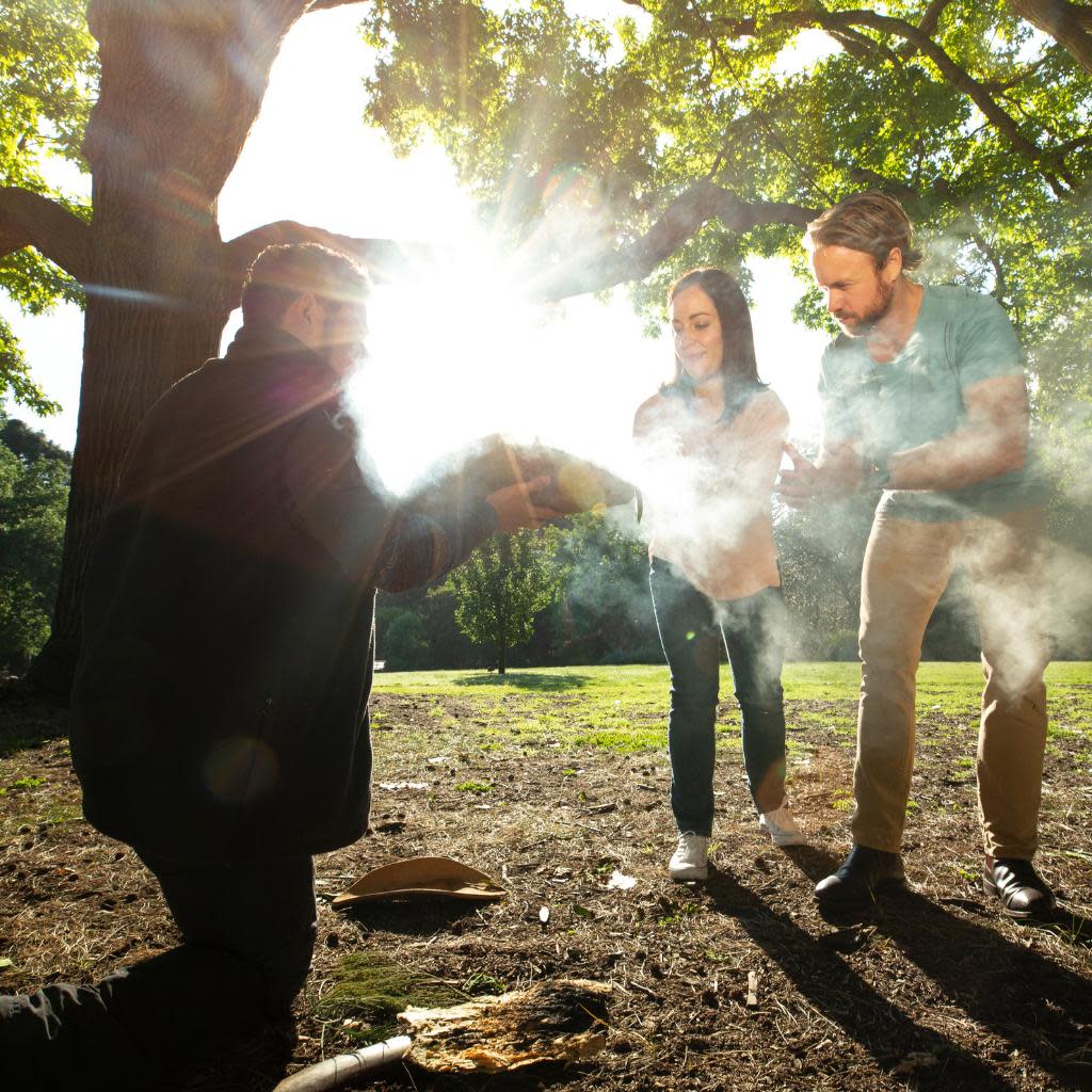 Aboriginal heritage walk in the Royal Botanic Gardens -a man is showing a smoking ceremony to two people standing up with the bright sun radiating behind them. 