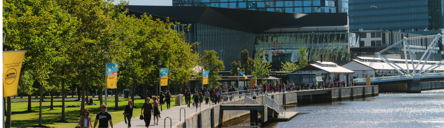 View down a boardwalk outside along the Yarra River. There are green trees running alongside the board walk and it angles toward a large building in the distance. 