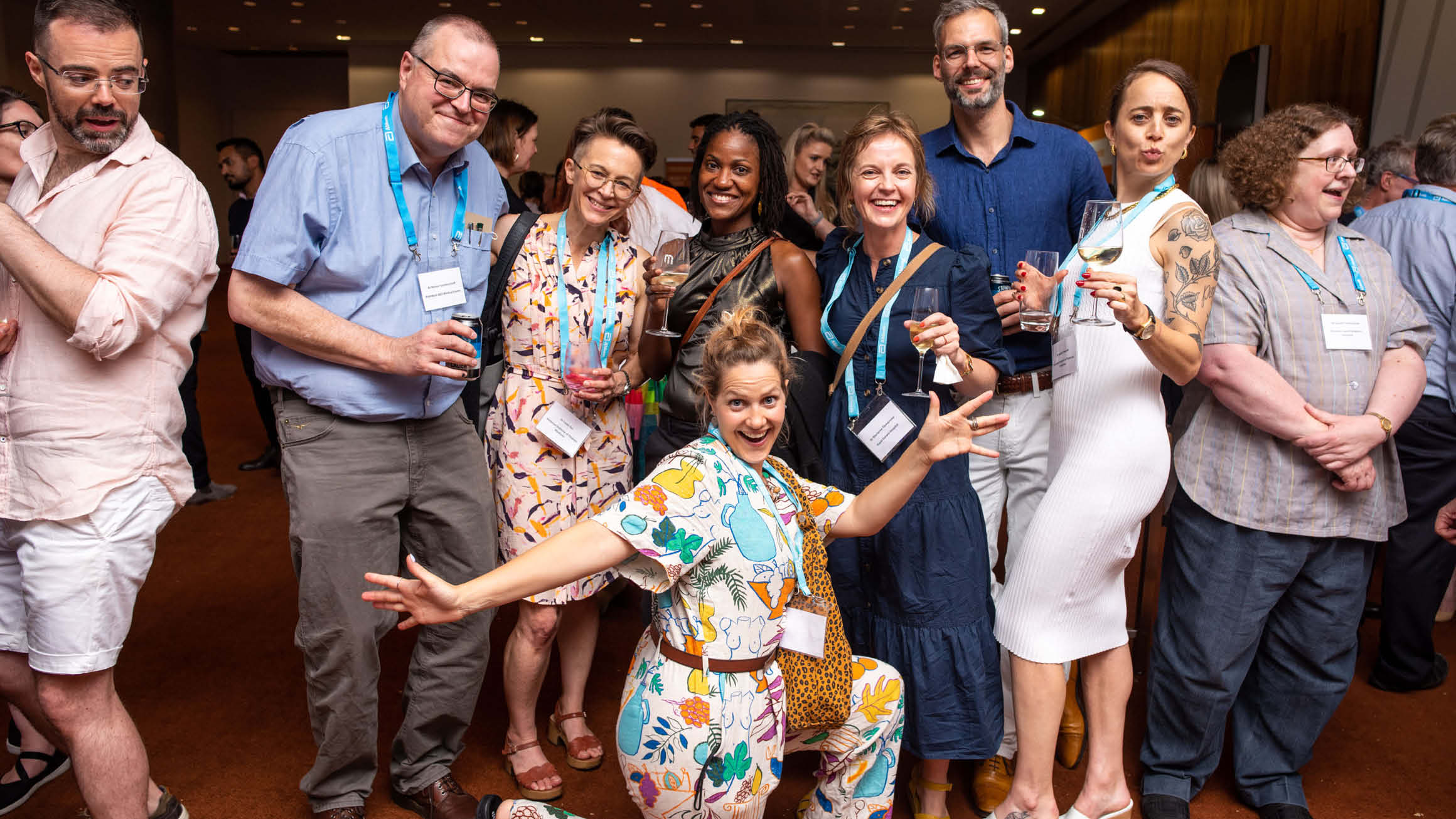 Group of people smiling at the camera all wearing badges to an event. Some are holding glasses of wine. 