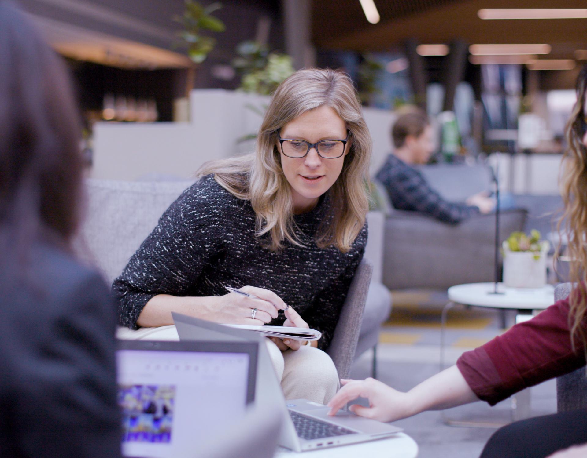 Woman sitting and leaning towards a computer on a table, in discussion with another person off screen. She wears a dark sweater and has her hair out. 