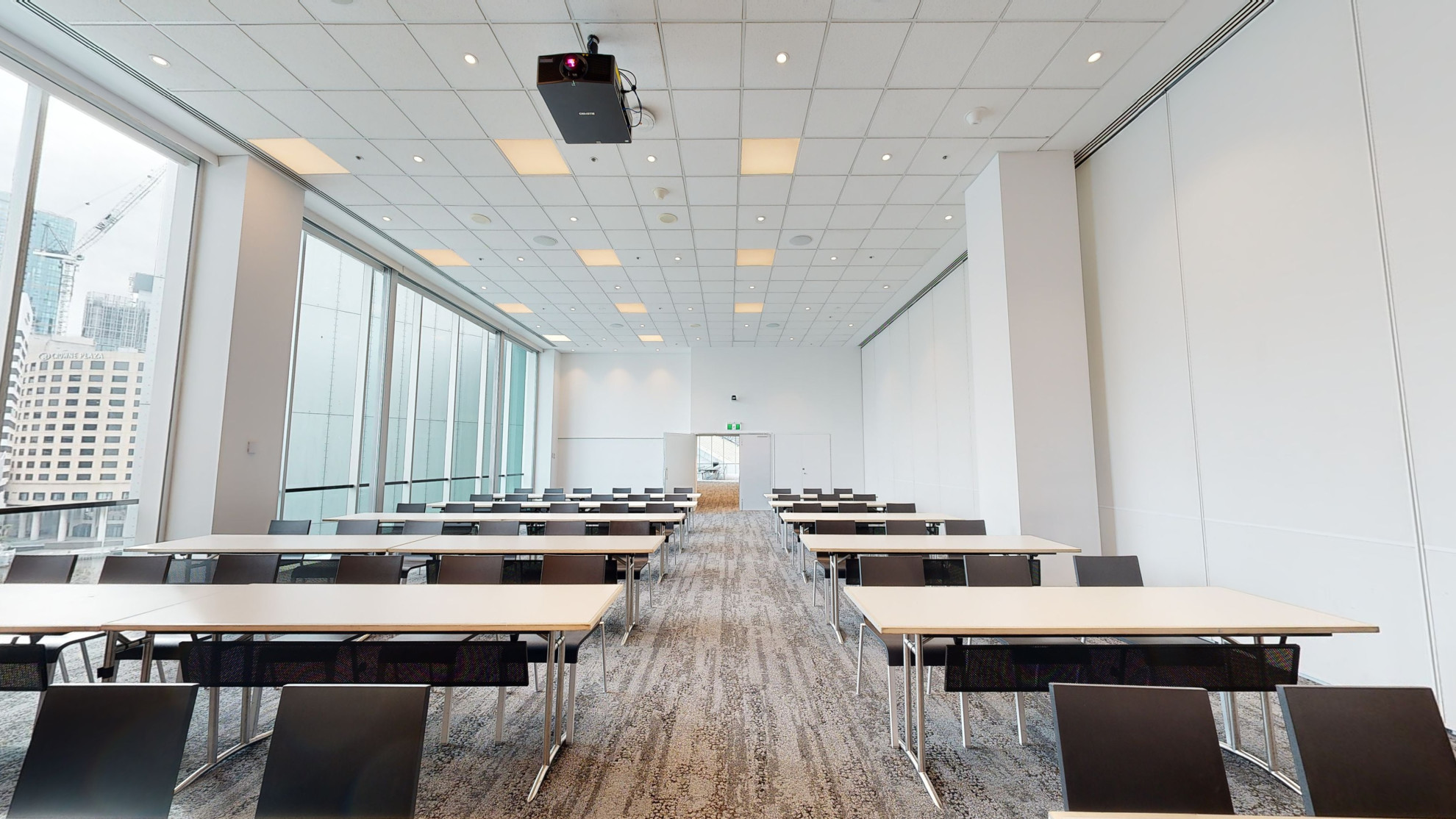 A professional conference or meeting room featuring a spacious layout, equipped with comfortable chairs for meetings and presentations. Table and chairs are set out in a classroom formation of rows. A large floor to ceiling window fills one wall of the room, which looks out to the Melbourne city. 