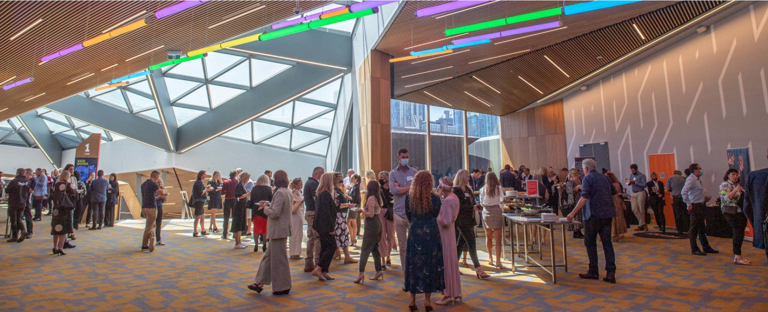 A lively foyer filled with a diverse crowd engaged in conversation and mingling. Overhead, vibrant bars of multi-coloured lights in shades of blue, purple, green, yellow, and orange hang from the ceiling.