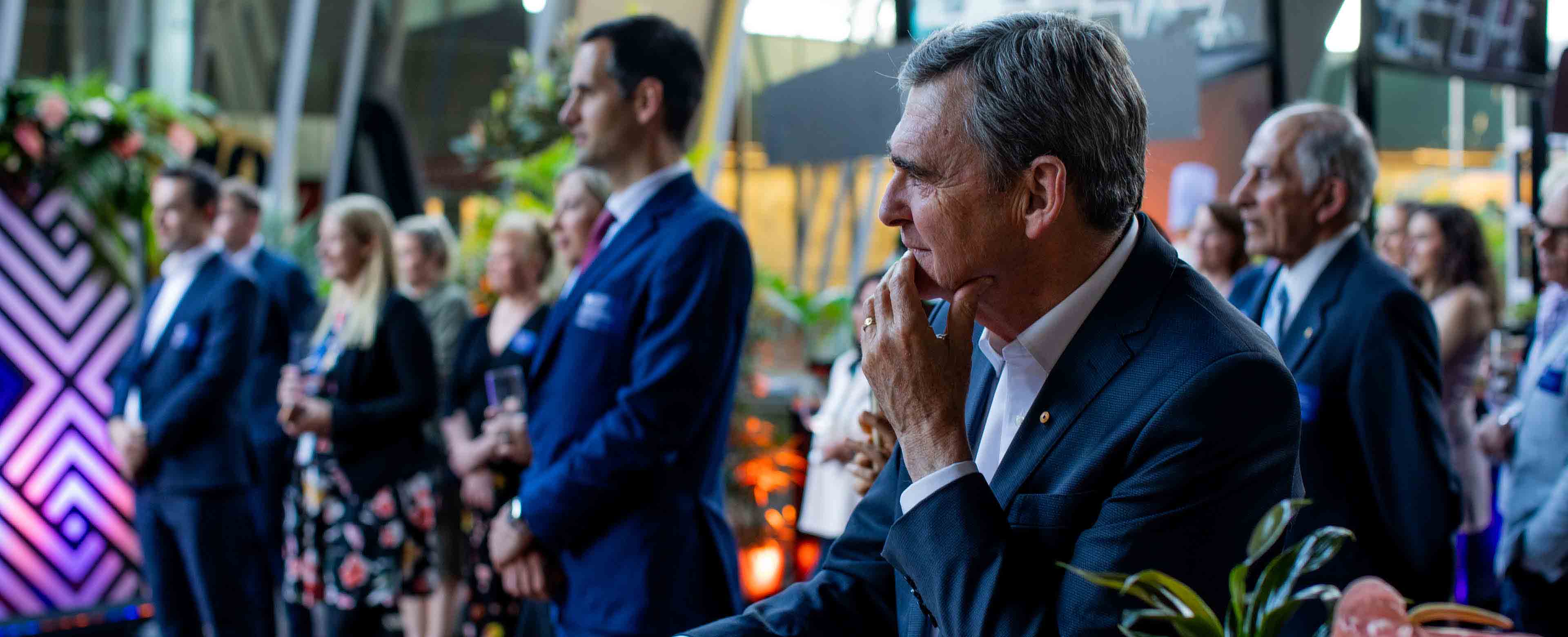 A man is sitting down and listening intently to a speaker. There are many other people in the background, also listening to the speaker. The man is looking thoughtful, as if he is considering what the speaker is saying.