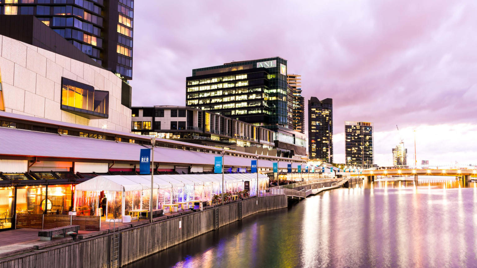 An image showcasing the vibrant South Wharf precinct in Melbourne. The Yarra/Birrarung River takes center stage, elegantly reflecting the pathway lined with stylish apartment buildings and restaurants.