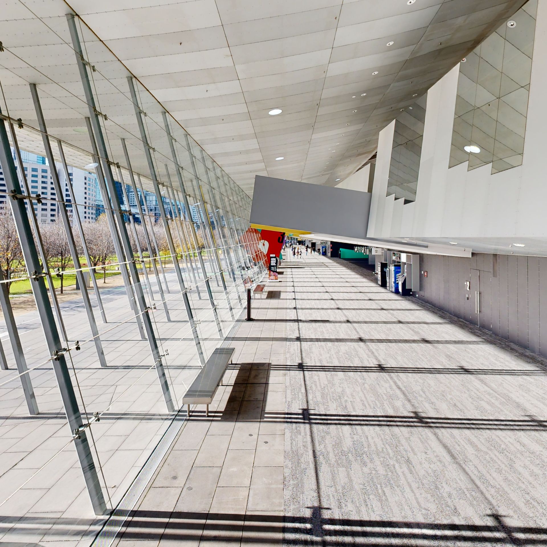 View of a large walkway with grey carpet. Large double story windows line one side with light streaming through. Metal poles that support the windows cast a grid layout of shadows onto the carpet. 