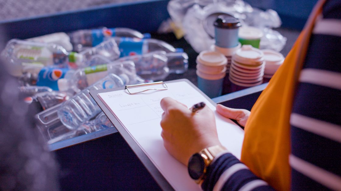 A person holding a clipboard, writing notes about recycled materials which lay across a table in front of them. 