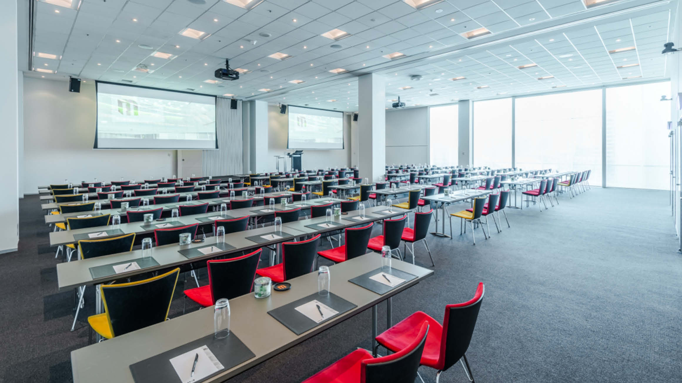 Empty room of tables and chairs lined in a classroom configuration. Chairs are facing the front of the room where two large screens are out. Floor to ceiling windows line the far wall letting in lots of natural light. 