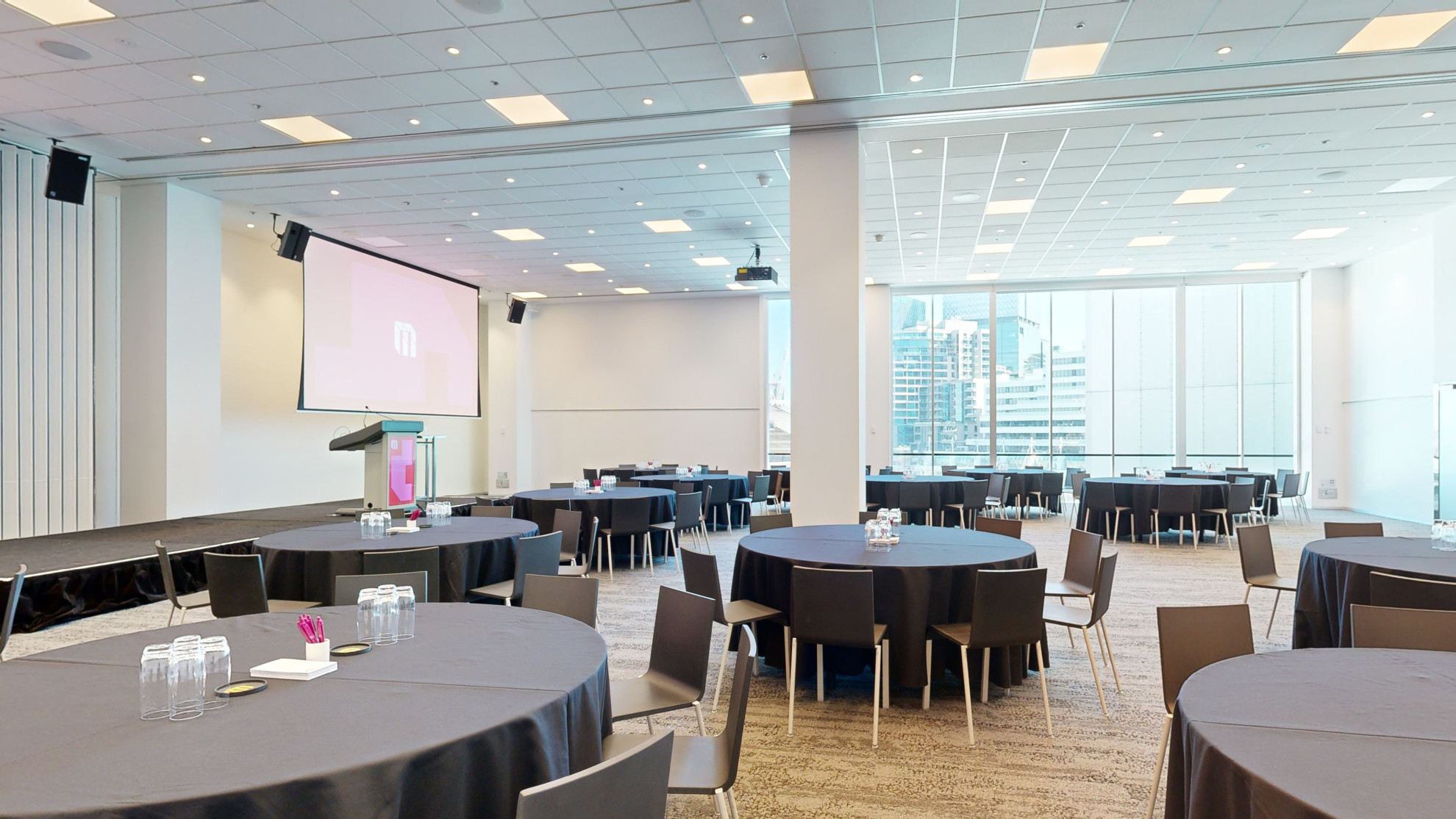 A spacious conference room with tables and chairs arranged for a meeting facing two large screens at the front of the room. Large floor to ceiling windows are to one side of the room. 