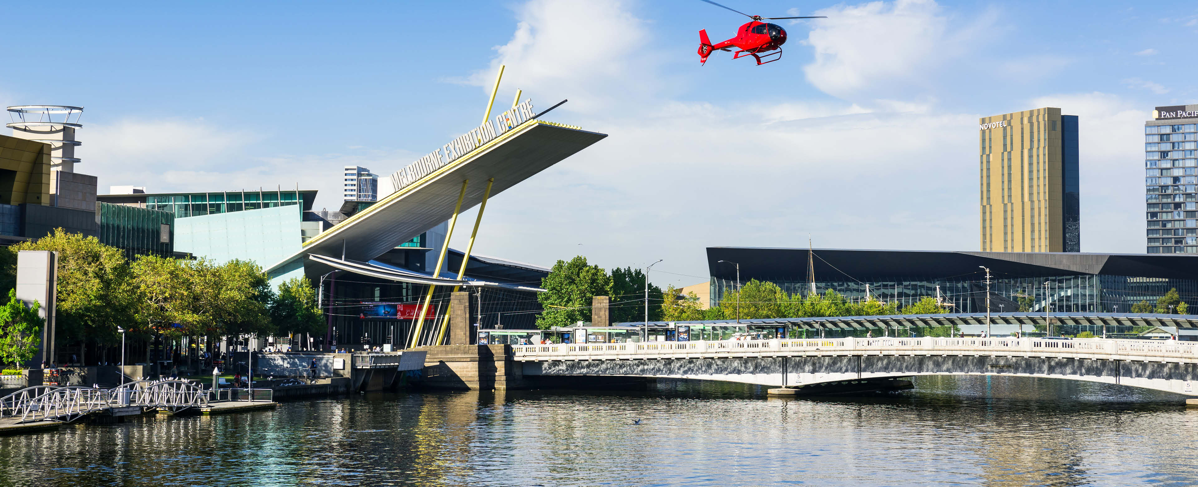 River view of the Exhibition building. A bridge extends over the river with a red helicopter flying in the blue sky. 