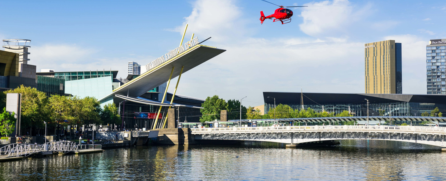 River view of the Exhibition building. A bridge extends over the river with a red helicopter flying in the blue sky. 