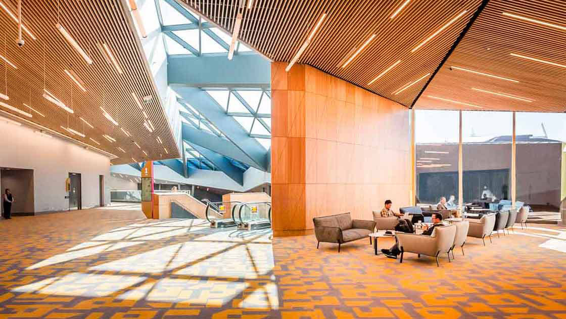 A bright orange foyer is filled with natural light from the floor-to-ceiling windows. A small group of people are sitting on lounge chairs, working on their phones, laptops, and books. The shadows from the people and the furniture dance on the floor, creating a dynamic and inviting space.
