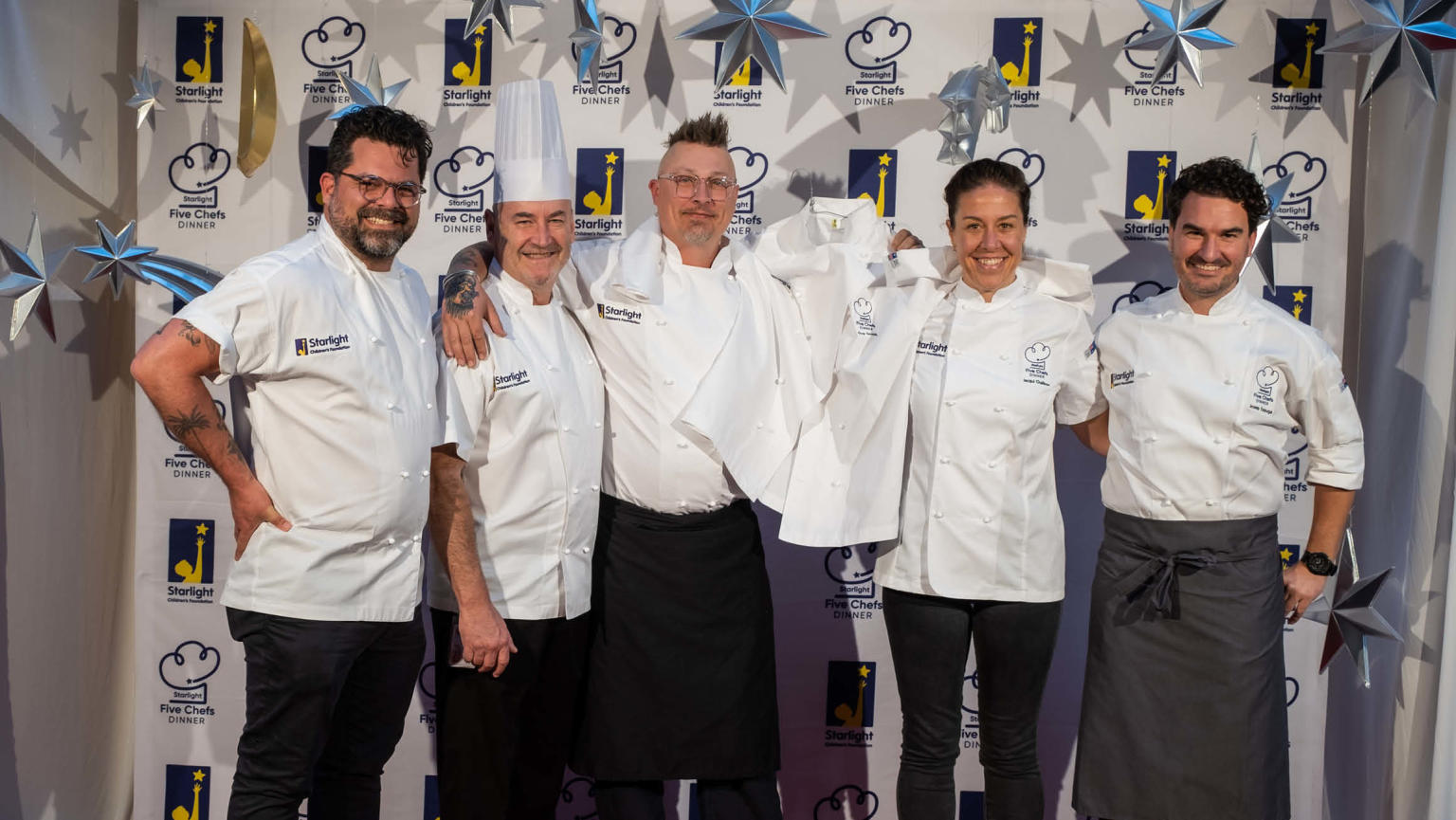 A lineup of five cheerful chefs, standing side by side and proudly holding up a crisp white shirt between two of them. They pose in front of a photo event backdrop, exuding a sense of camaraderie and professional pride.