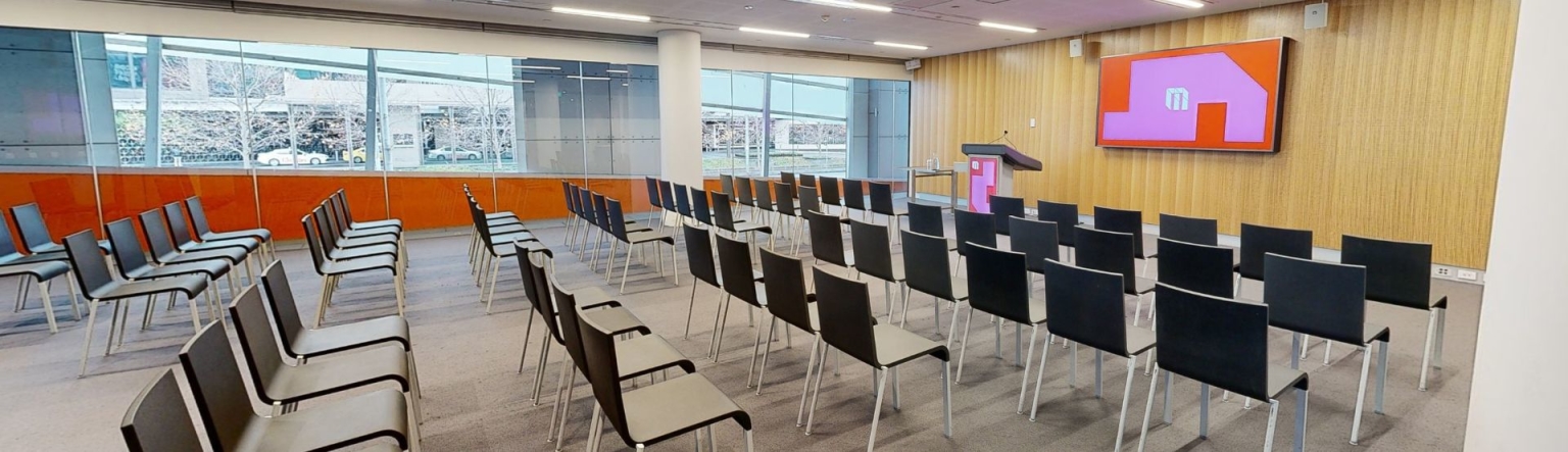 A conference or meeting room featuring rows of chairs arranged to face large tv screen for presentations and discussions. A lectern sits to the side of the tv screen and windows run along the side of the room facing out towards Crown. 