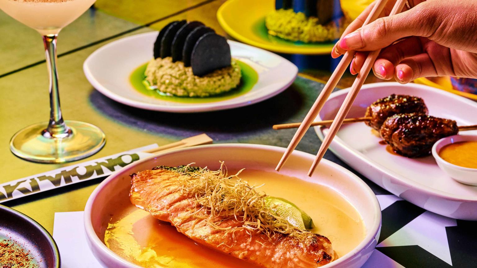 Bright table with lots of food plates. A hand is holding chop sticks over one of the plates. 