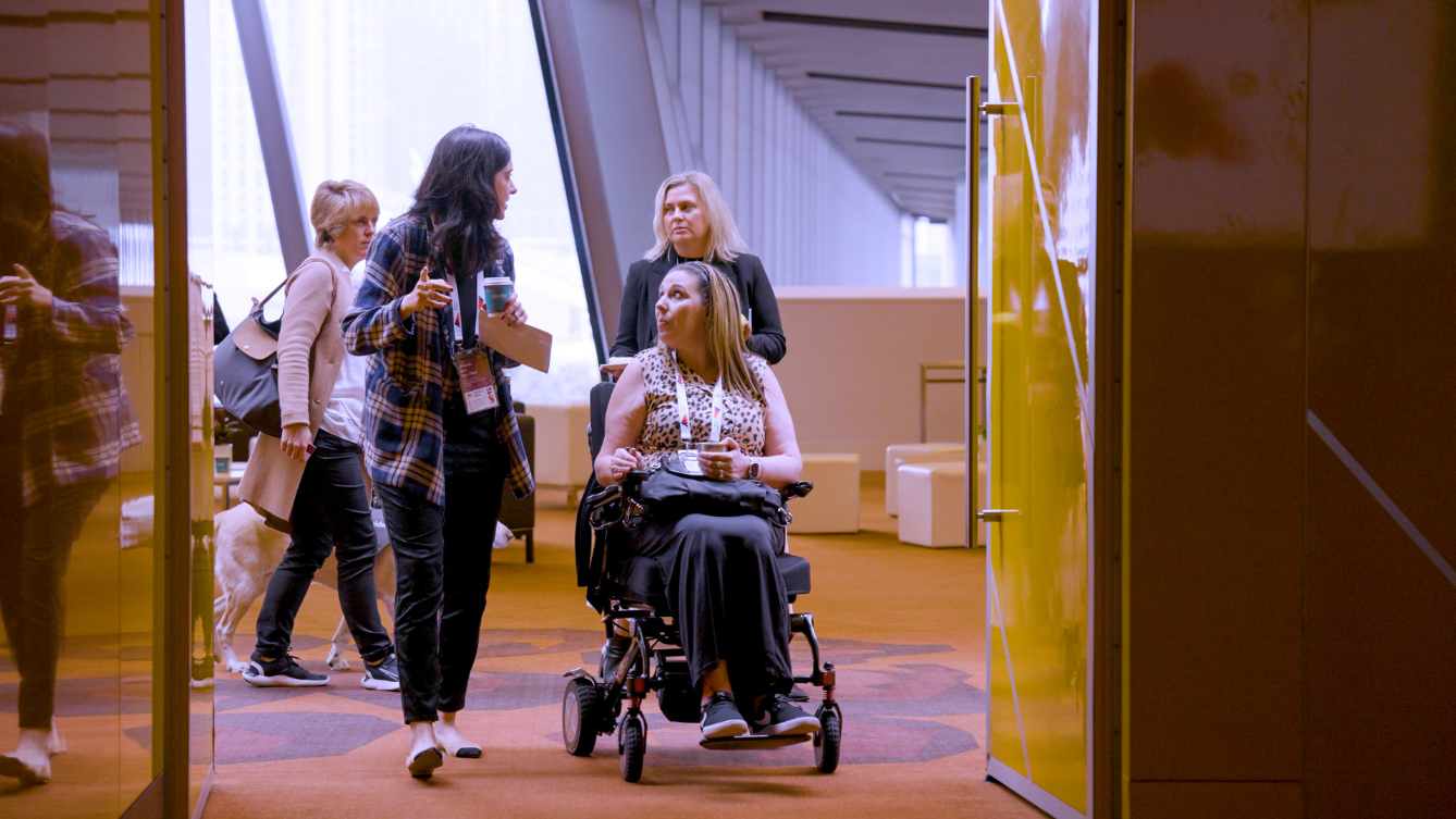 Two women are entering a room, a large yellow double door is opened for them to enter through. One lady is in a mobility scooter while the other walks beside her holding a coffee. 