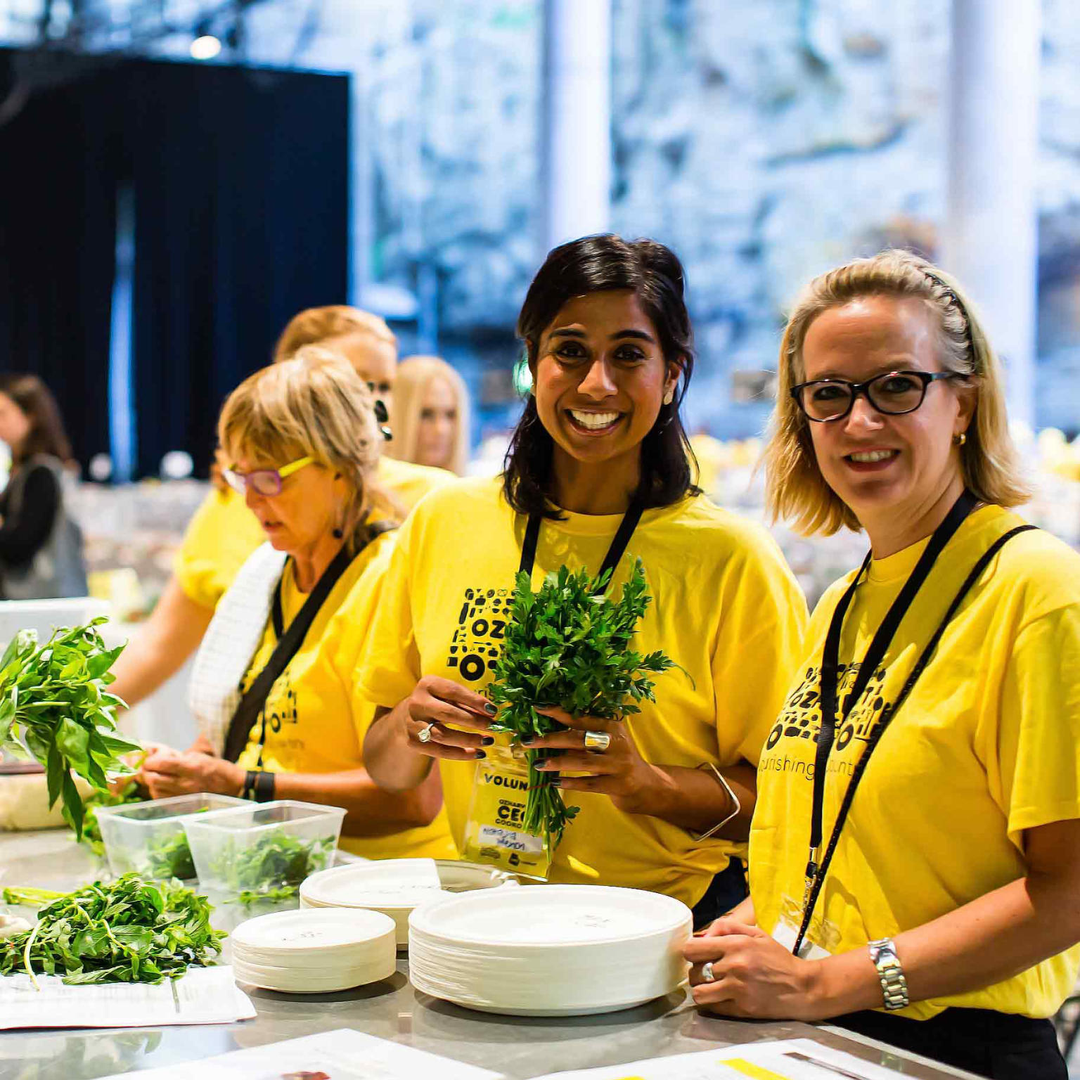 Two woman smile at camera wearing yellow t-shirts. They are preparing food at a table. One woman is holding a bunch of green herbs. More people are in the background also wearing yellow t-shirts. 