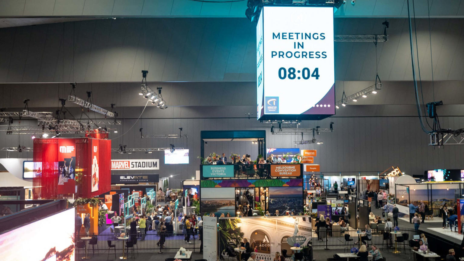 A spacious exhibition area showcasing various stands and bustling with attendees. In the centre hangs a prominent digital sign displaying the message 'Meeting in progress 8:04,' while numerous individuals are seen walking around.