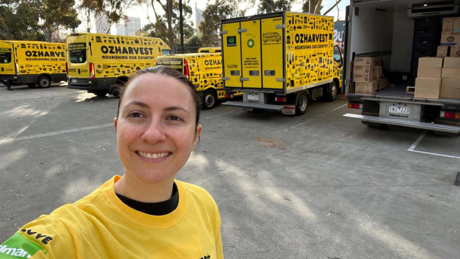 An image showing an MCEC employee wearing a yellow OzHarvest t-shirt taking a selfie of her smiling in front of various yellow OzHarvest vans. 