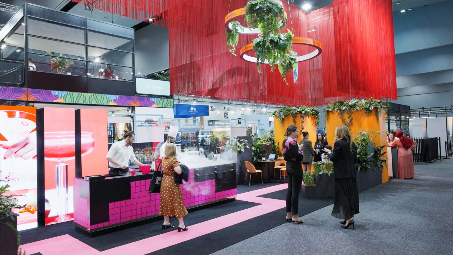 An eye-catching exhibition stand featuring various elements: long red fringing suspended from the ceiling, orange circles adorned with hanging plants, a black and pink table where a man in a white shirt serves ice cream, a captivating digital screen displaying a cocktail in a coupe glass. The stand is enhanced by black and pink carpeting, and three individuals standing nearby.