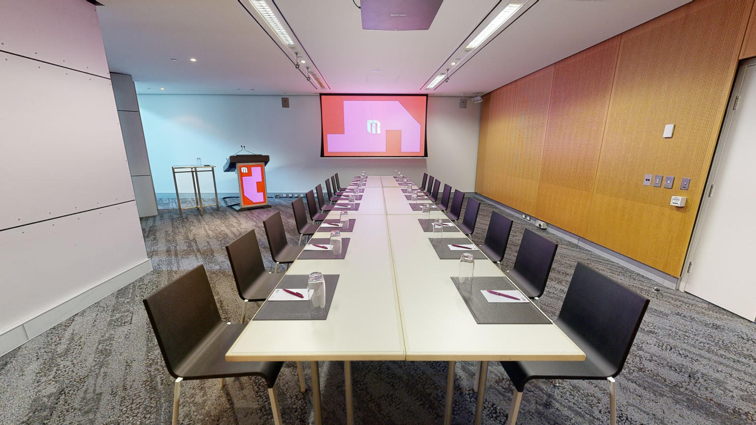 A conference or meeting room featuring a spacious boardroom layout equipped with a sizable screen and comfortable chairs for attendees. A lectern also sits in the corner to the left of the screen.