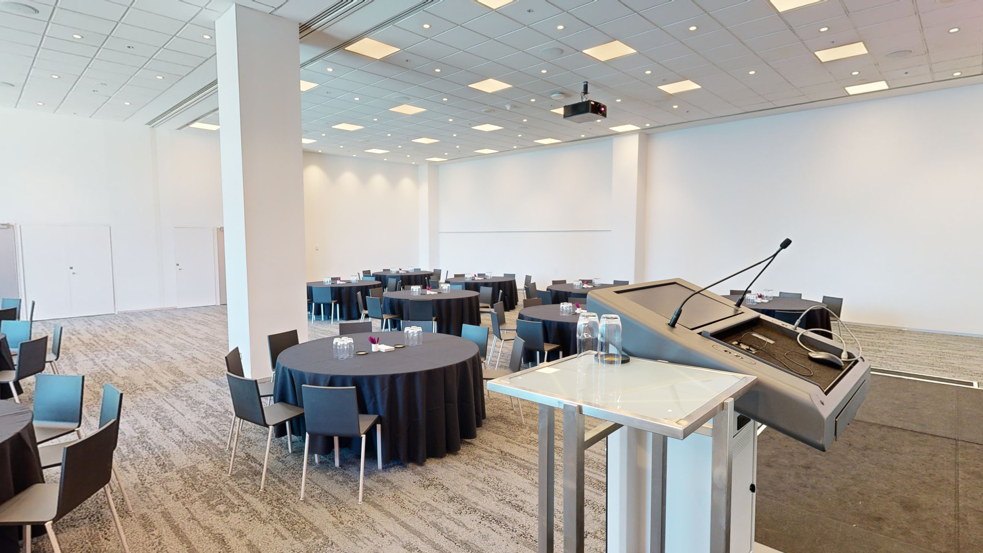 View from a lectern at the front of the room facing a spacious room with tables and chairs. Large floor to ceiling windows are to one side of the room. 