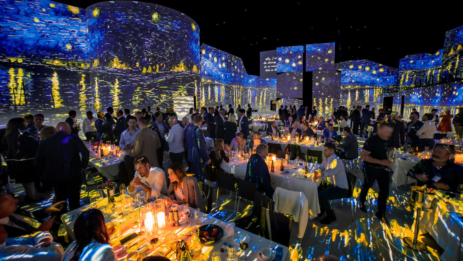 Captivating gala event with guests dining at long tables, surrounded by immersive digital screens showcasing Van Gogh's iconic 'Starry Night' artwork. The mesmerising reflection is featured on the floor and throughout the venue.