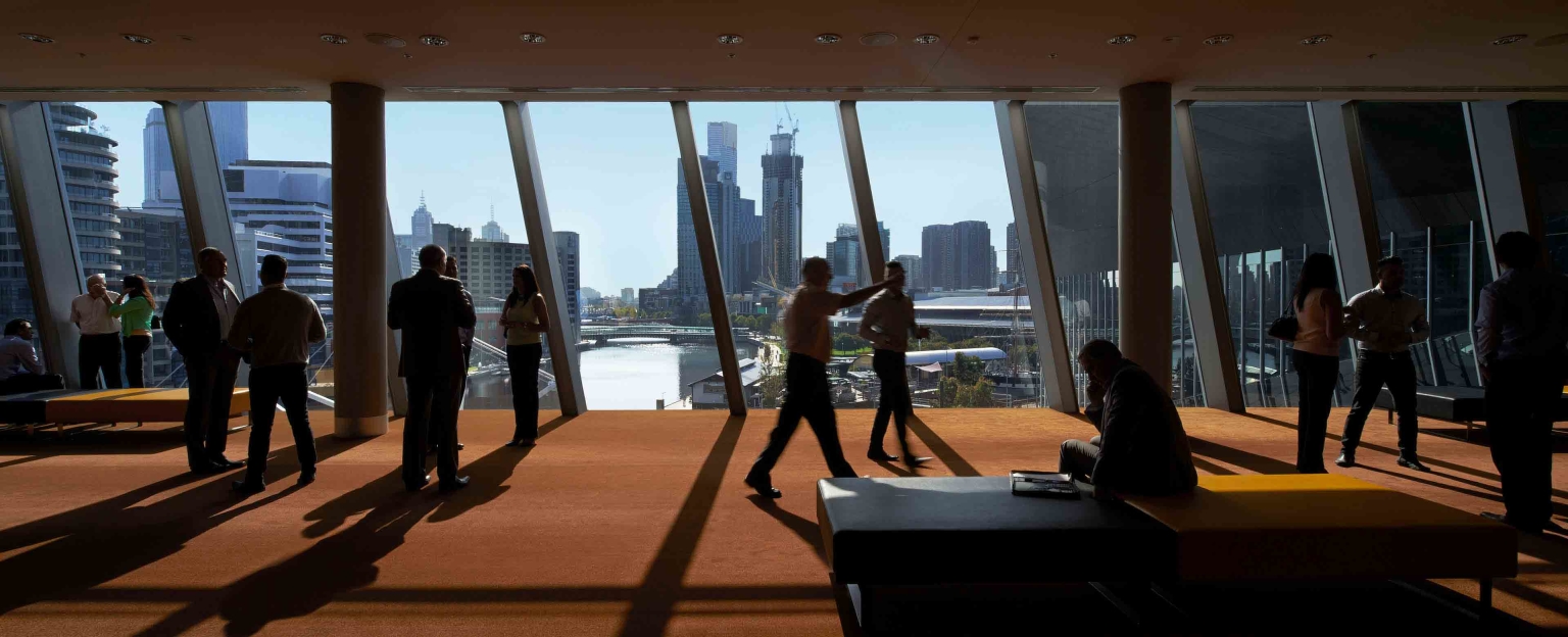 A group of people walk through the foyer of the Melbourne Room at Melbourne Convention and Exhibition Centre. The foyer has a large window that looks out onto the Yarra/Birrarung River. The city skyline is visible in the background.