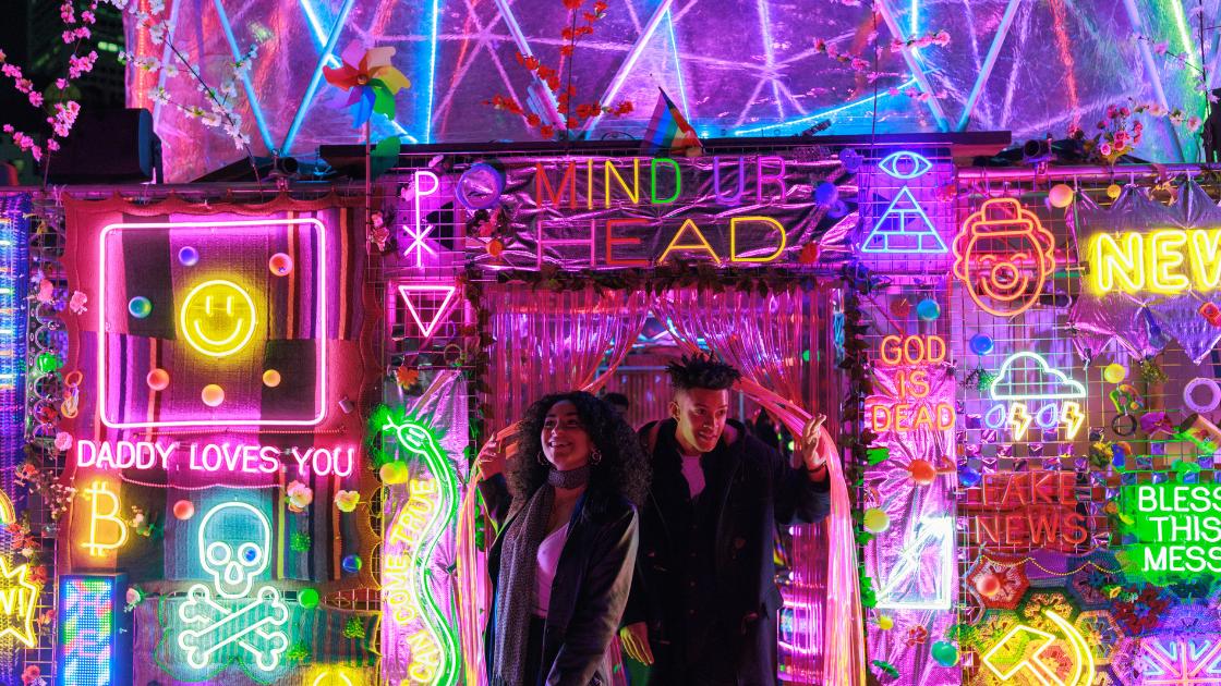 Two people walk through a doorway to a brightly lit room of neon signs. 