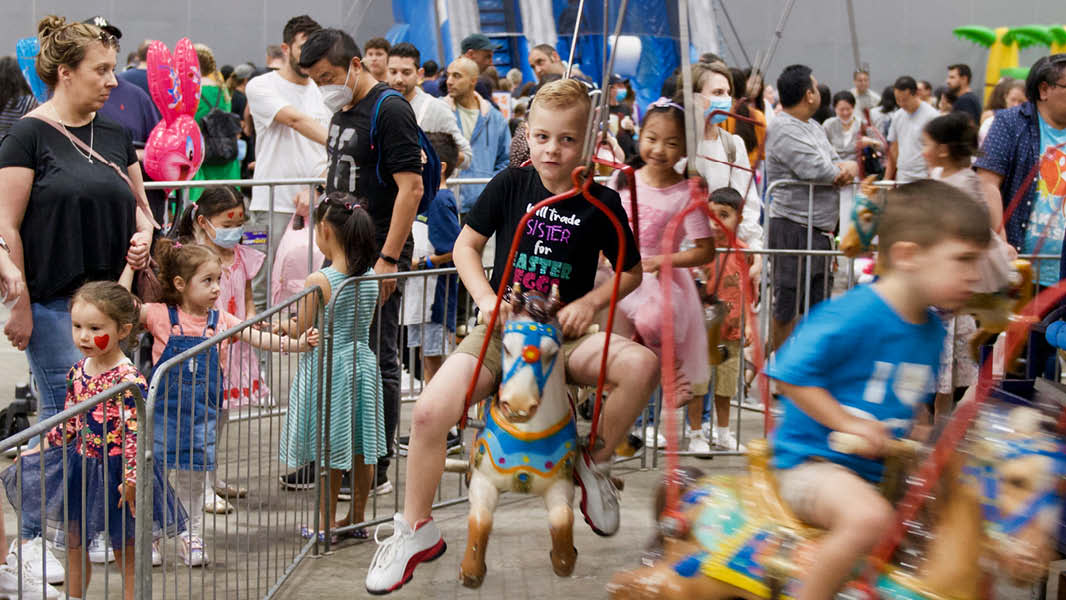 Children sitting on a horse carousel with a busy exhibition of people walking around in the background. 
