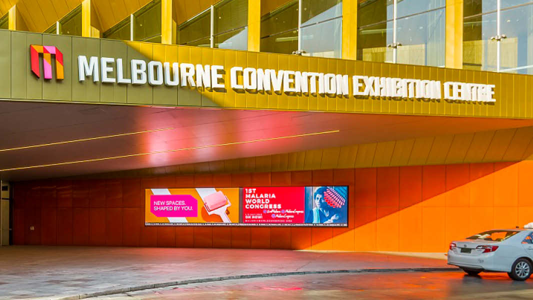 A large gold building featuring digital signage on the entrance wall, showcasing vibrant displays. A prominent logo is positioned on a protruding block, proudly displaying the words "Melbourne Convention Exhibition Centre."