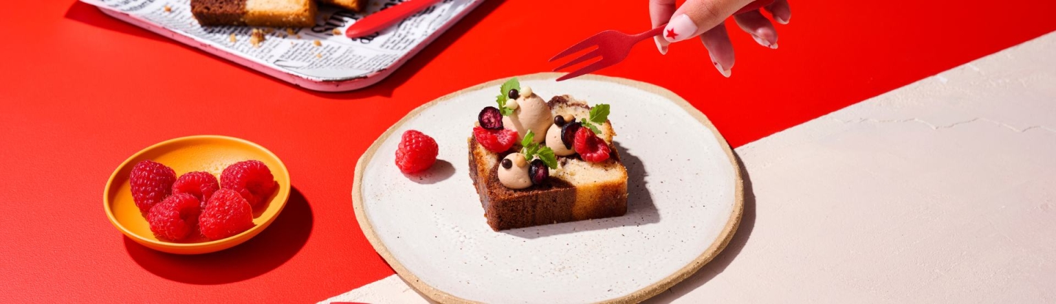 Chocolate, vanilla and coffee marble cake presented on a white plate. The plate sits on top of a red table top with an orange bowl to the side full of raspberries. 