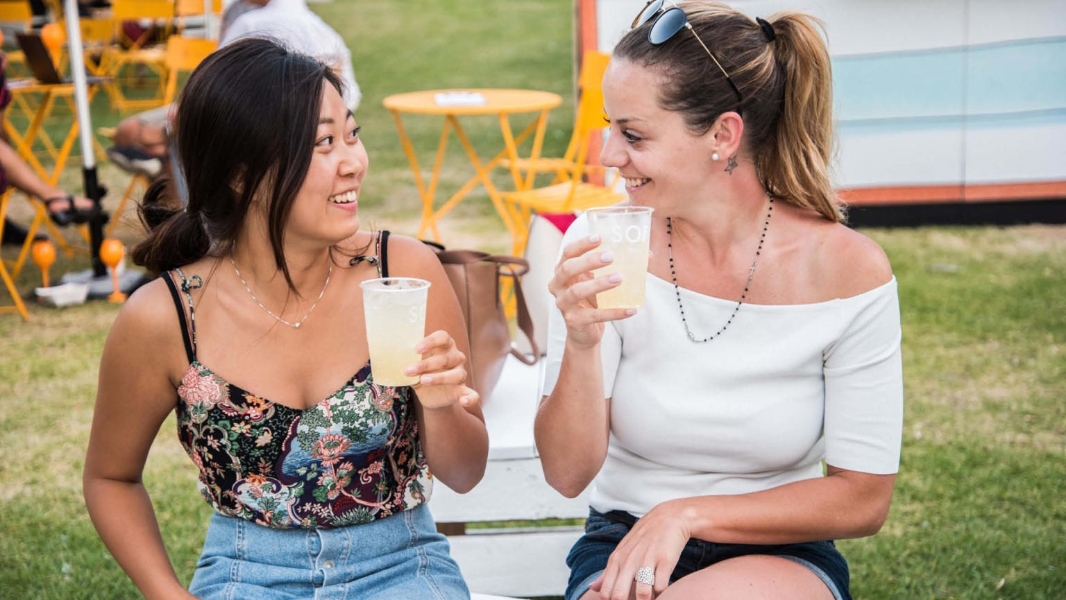 Two women seated outdoors, sharing a joyful moment as they gaze at each other with smiles, each holding a delightful light yellow beverage.