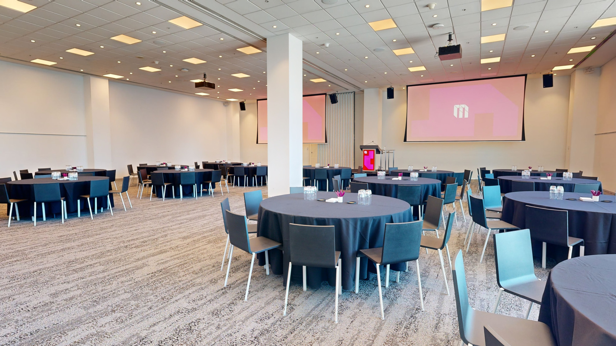 A spacious conference room with tables and chairs arranged for a meeting facing a podium with a lectern at the front of the room. Large floor to ceiling windows are to one side of the room. 