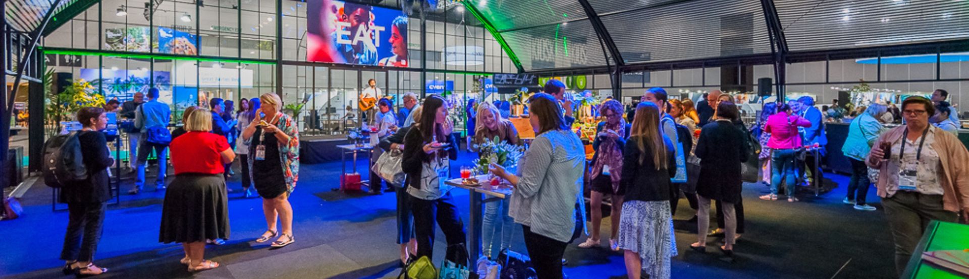 Large crowd of people at an exhibition, standing around standing tables talking, with blue and green lighting projected throughout. 