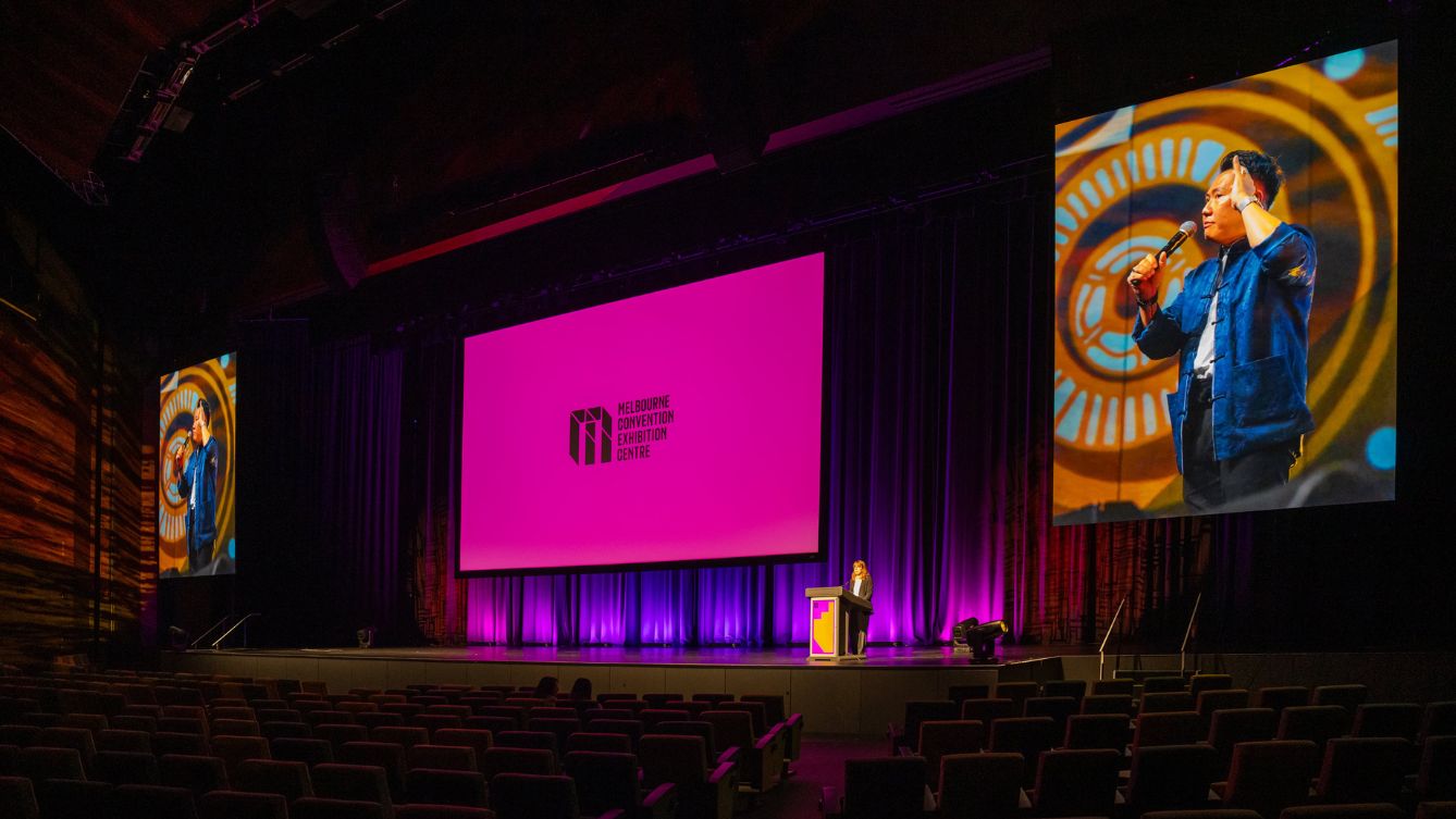 Large drop down screen with the MCEC logo on a pink background. Two other screens, one of either side displaying a person talking into a microphone. 