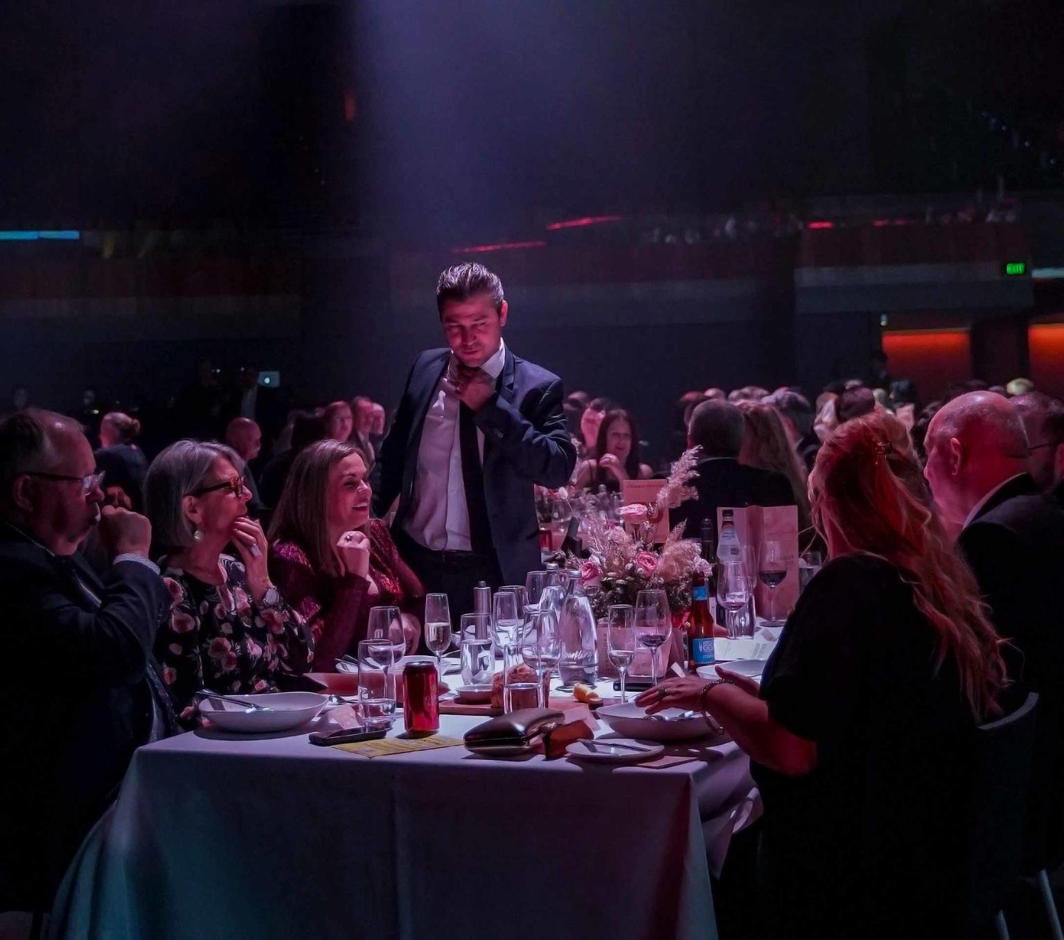 A man standing in front of a crowd of people who are seated at a gala dinner. 
