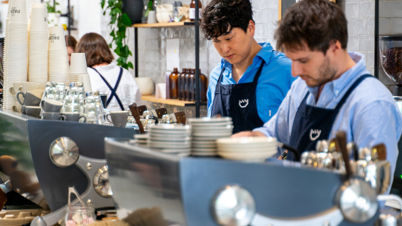 Two guys behind a coffee machine, making coffees. They are in a Padre uniform in what appears to be a cafe working. 